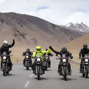Motorcyclists on a mountain road, some raising fists in triumph, with a snow-capped peak in the background in leh and ladakh.